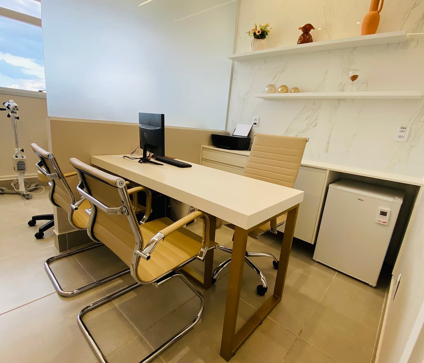 Consultório da unidade Prado do Instituto Hiraki com mesa de atendimento, cadeiras, frigobar e foco clínico